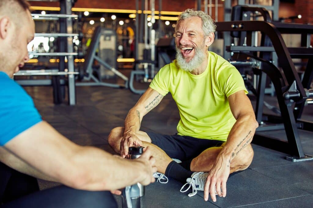 Cheerful middle aged man holding bottle of water, discussing something with fitness instructor or